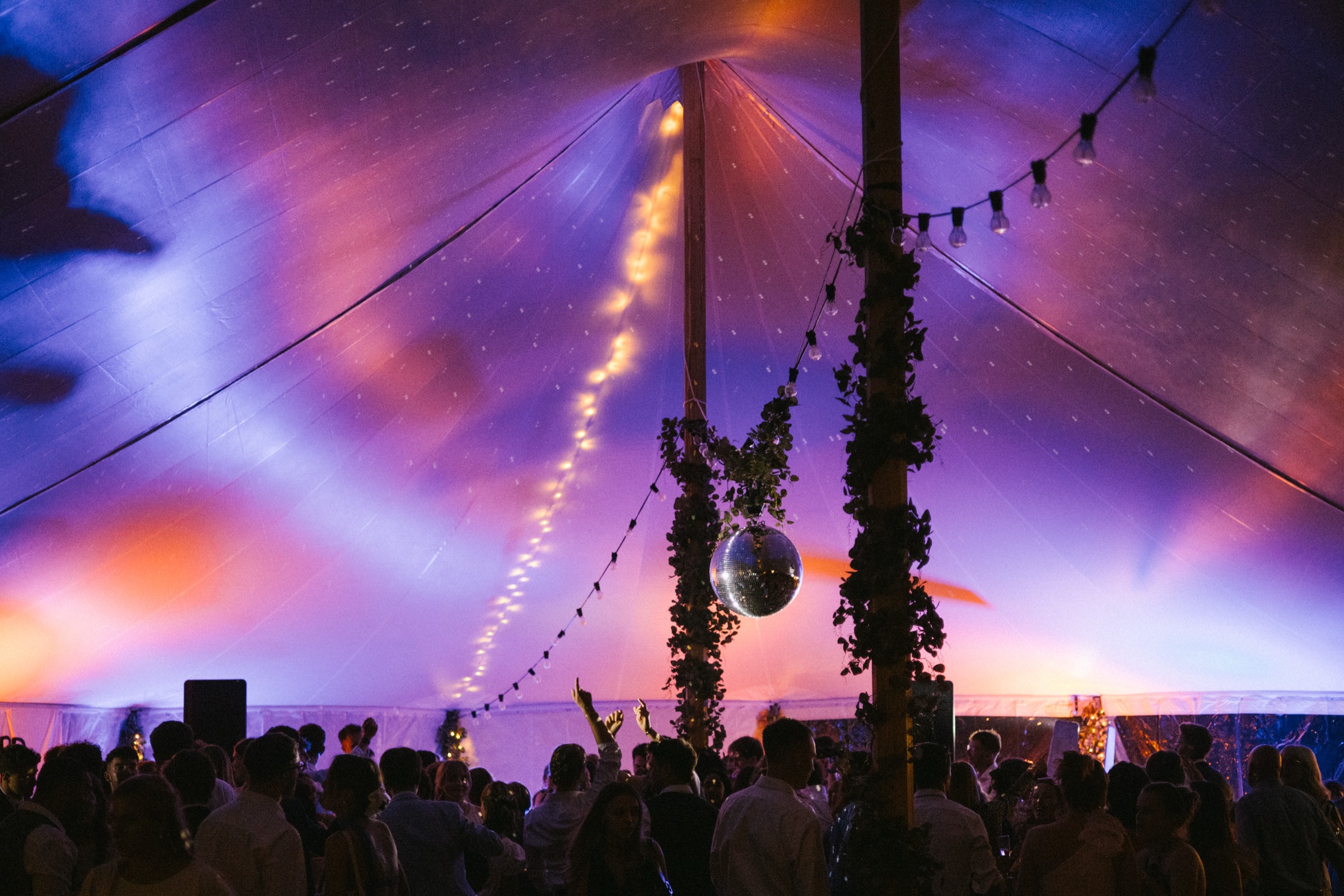 A packed wedding dancefloor under a glowing marquee with a disco ball and festoon lights